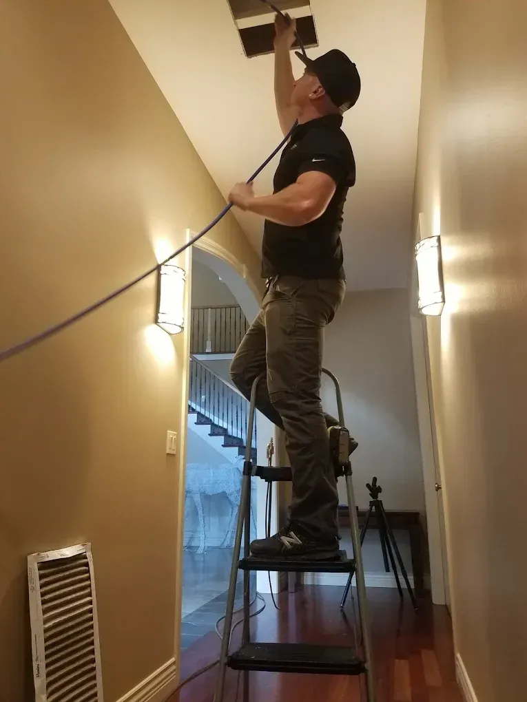 Man on stepladder cleaning air vent in hallway, wearing black shirt, dark pants, and cap.