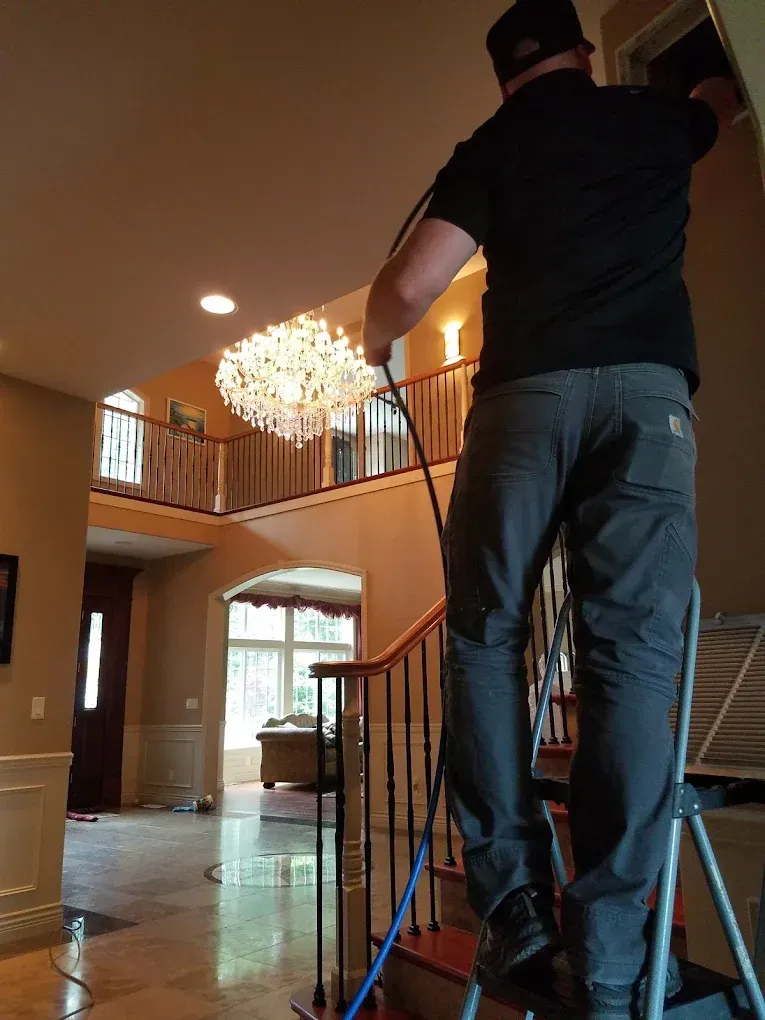 Man on ladder cleaning an air vent. Light-filled entryway with chandelier and a staircase.