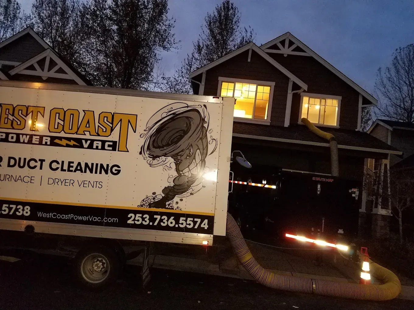 A duct cleaning truck parked outside a house at dusk, with a hose leading to the upper windows.
