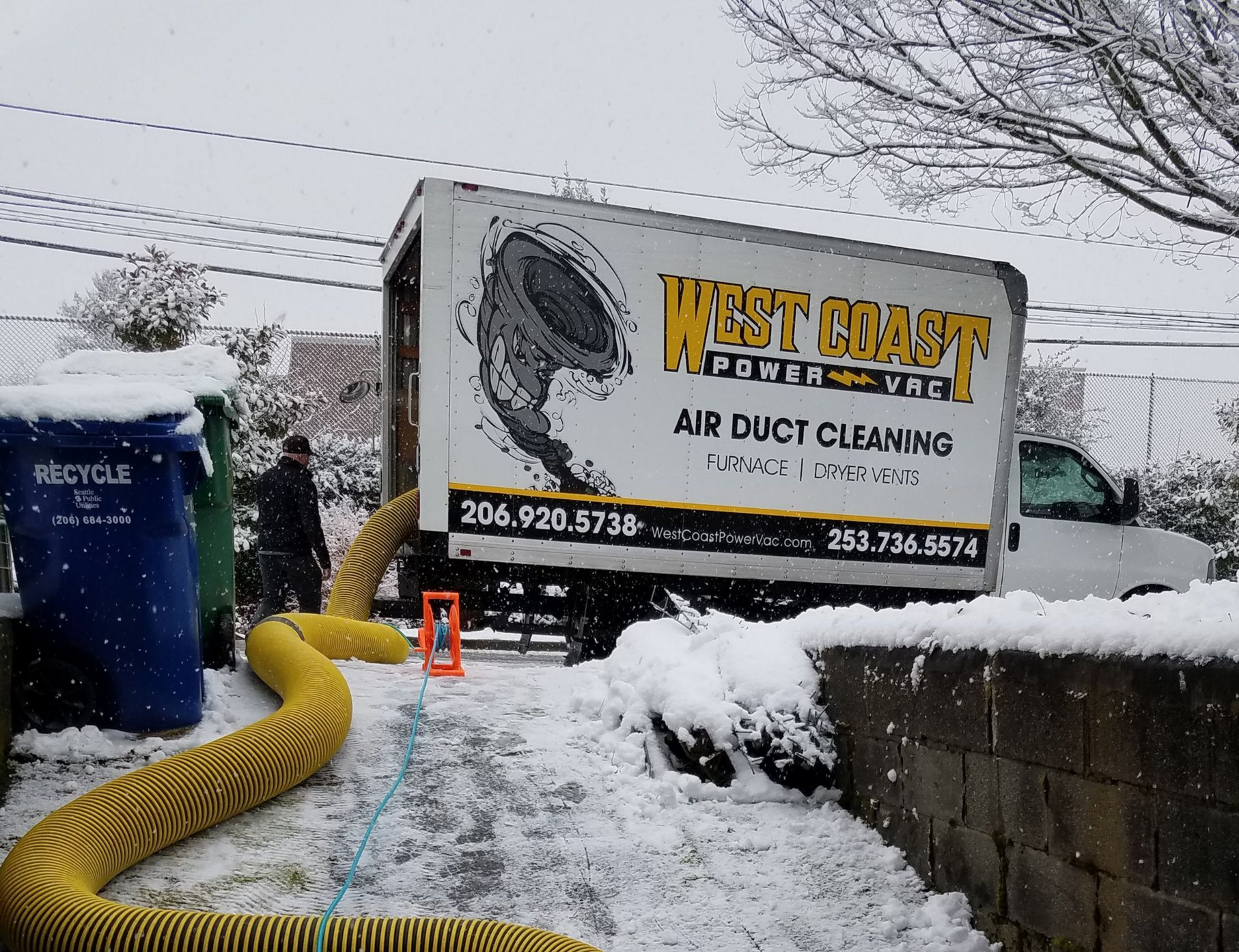 A West Coast Power Vac truck cleans air ducts in a snowy driveway. A yellow hose extends from the truck.
