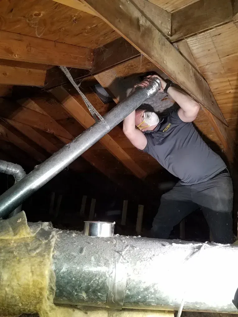 Man in an attic installing HVAC ductwork. He's wearing a mask and dark clothing. The space is dark.
