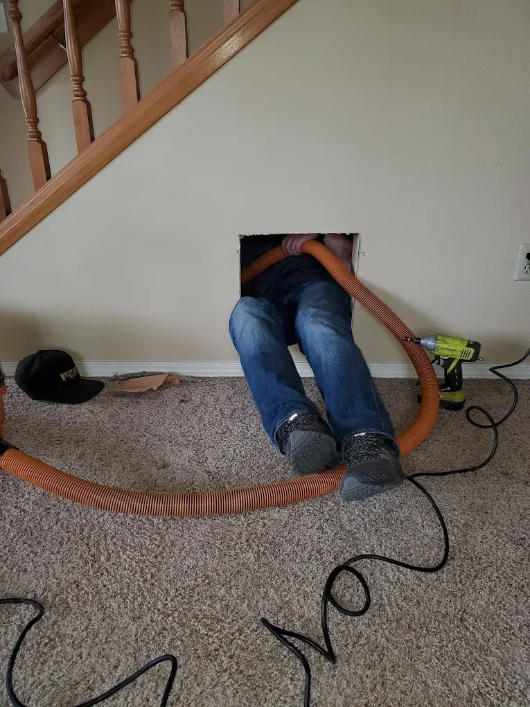 Person in jeans, leaning into a wall opening, holding a long orange hose, near stairs, vacuuming.
