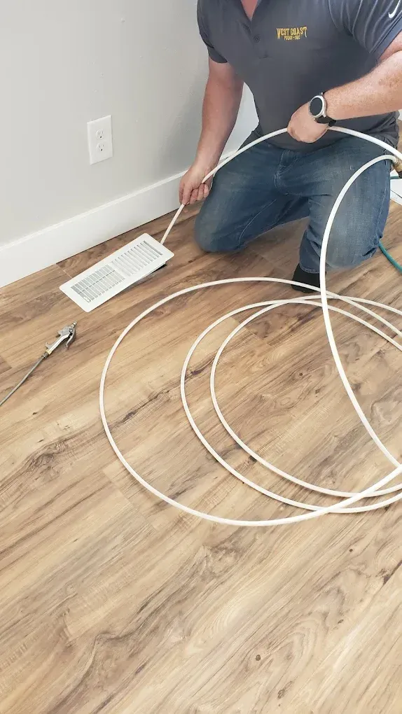 Man cleaning HVAC vent on wood floor. He is kneeling, using a white cable.