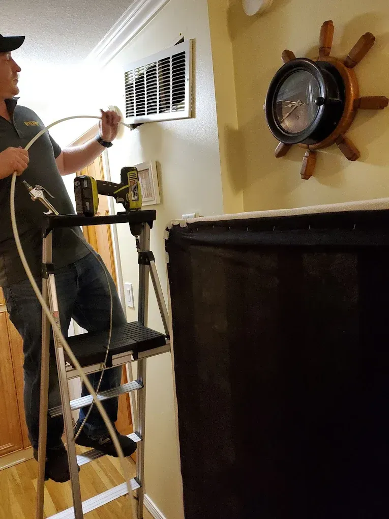 Man on ladder cleaning an air vent. He's using a hose, with a drill nearby. Nautical clock on the wall.