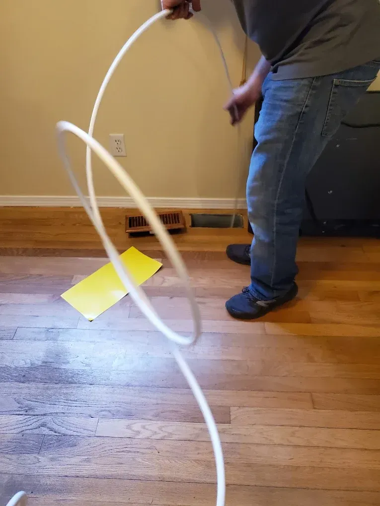 Person uncoils white tubing on wood floor near vent and electrical outlet.
