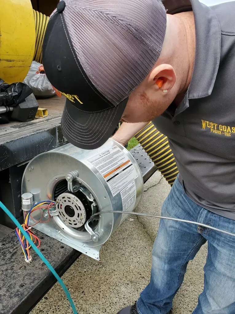 Man in gray shirt and cap, inspecting a silver blower fan outdoors.