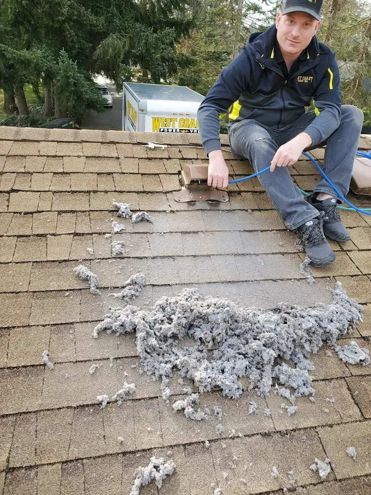 Man on a roof, removing a large pile of gray debris. Overcast day.