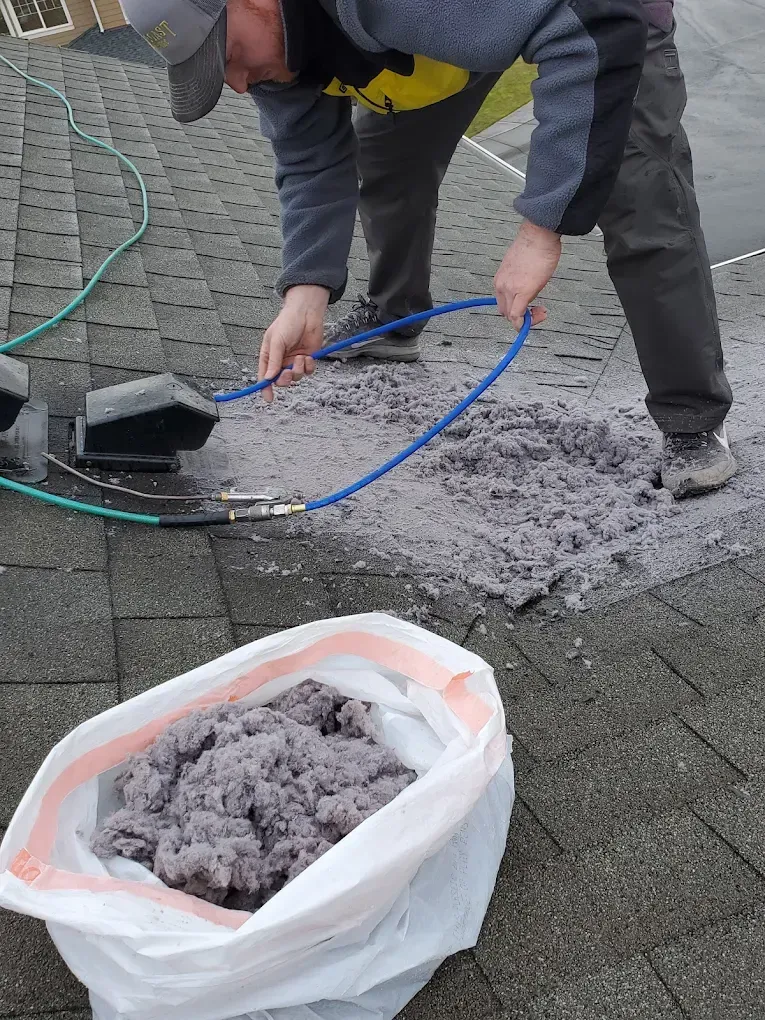 Man cleaning a roof vent with a hose, pile of gray lint nearby, bag of collected lint.