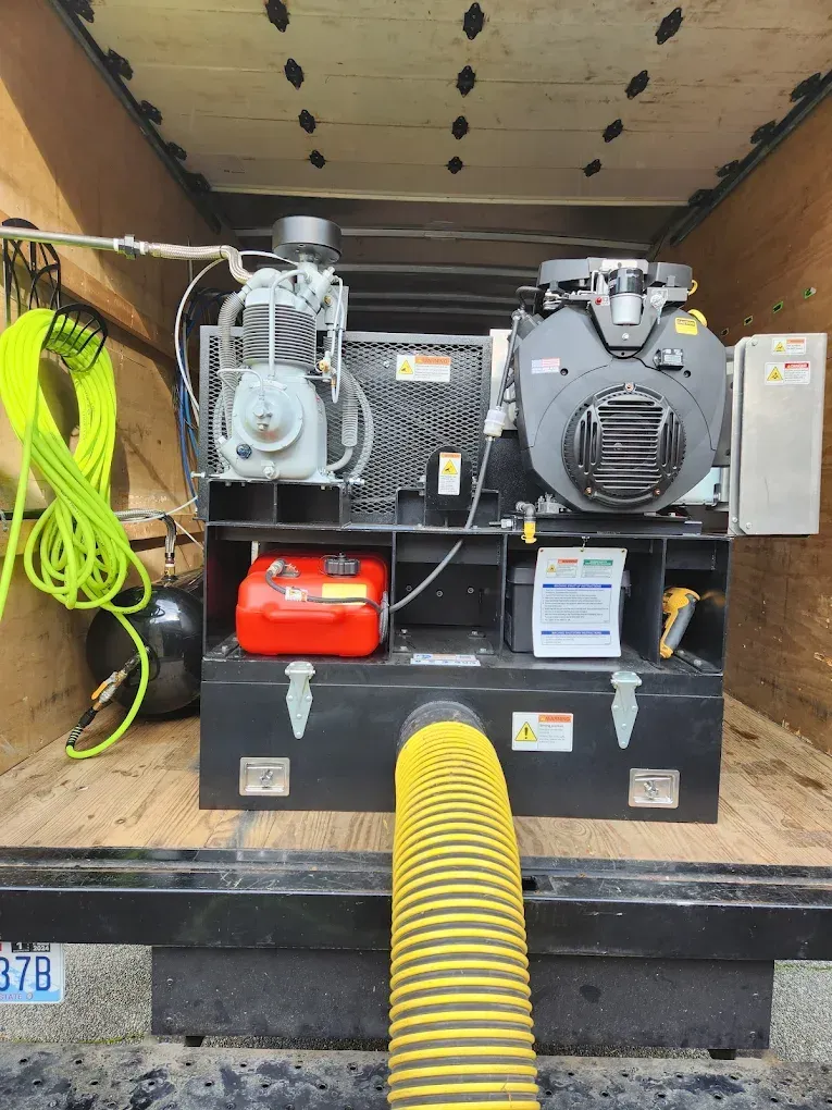 Black industrial machine inside a truck bed. Features a gas engine, compressor, fuel tank, and yellow hose.