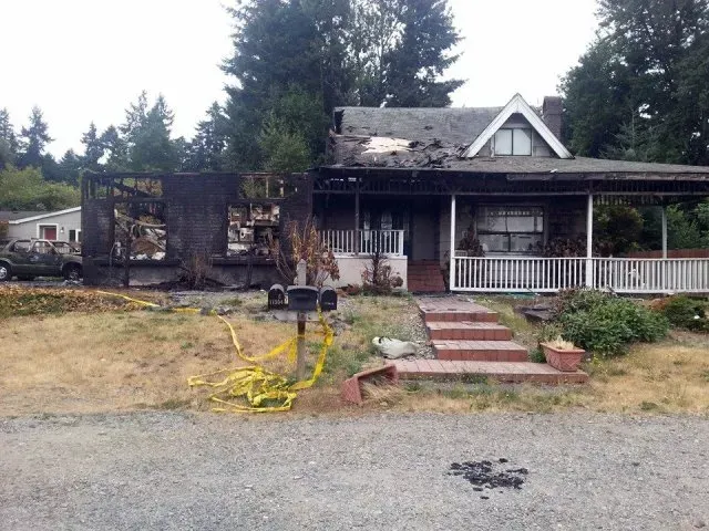 House damaged by fire; charred walls and roof, porch intact, yellow caution tape in front.