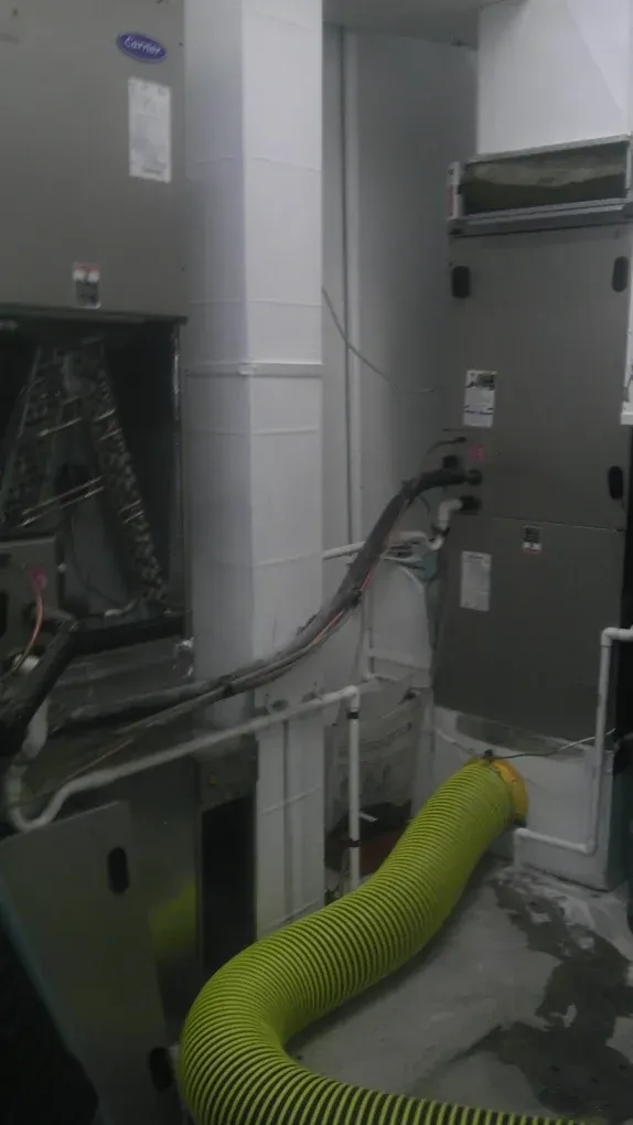 HVAC unit, grey metal, with black and yellow flexible ductwork, against a white wall.