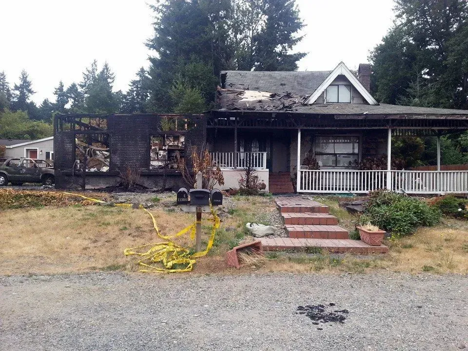 House with fire damage; charred walls, roof partially collapsed. Yellow caution tape on lawn.