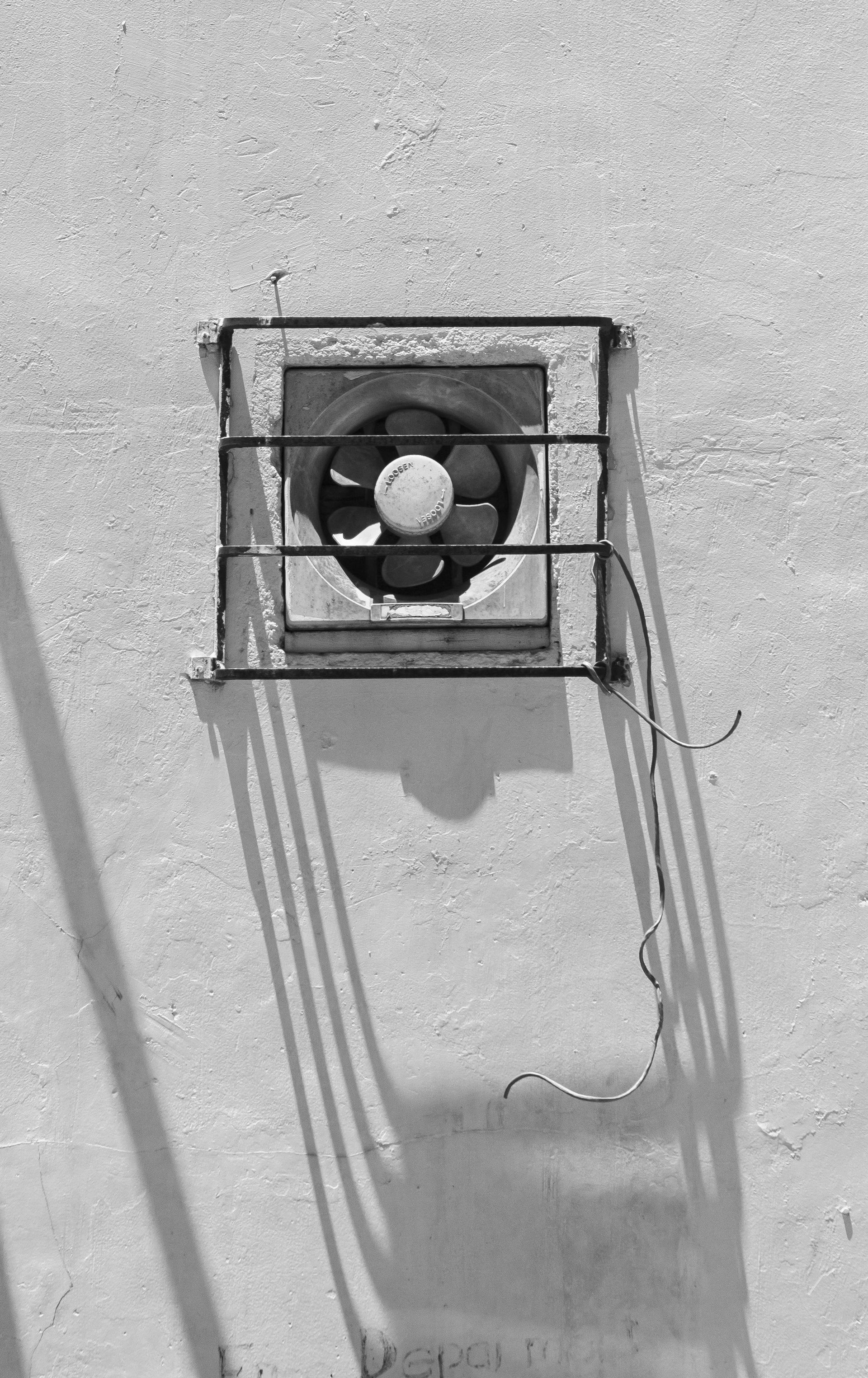 A black and white image of a ventilation fan on a stucco wall, with a security grill and casting a long shadow.