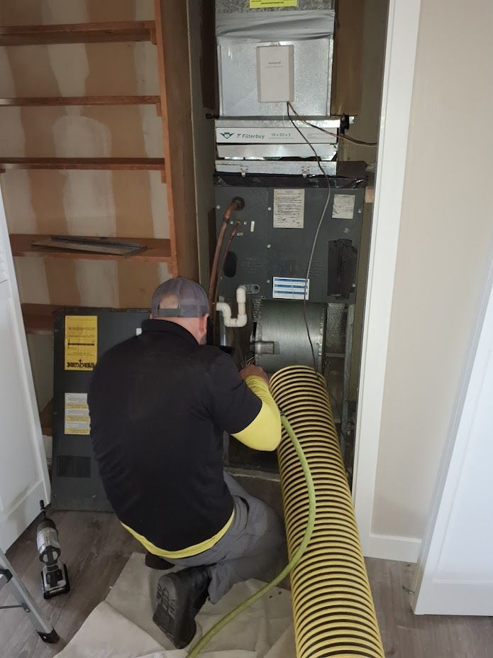 A man kneels, working on a furnace in a garage. He wears a gray shirt and black pants.