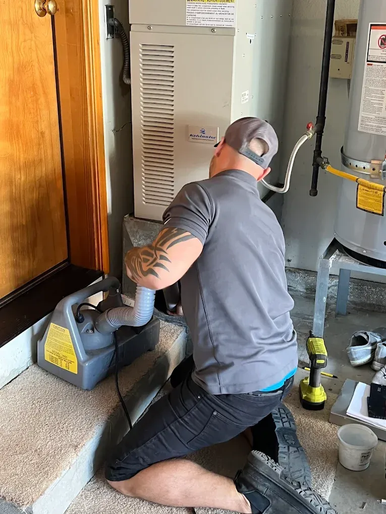 Man working on furnace in garage, wearing grey shirt and baseball cap.