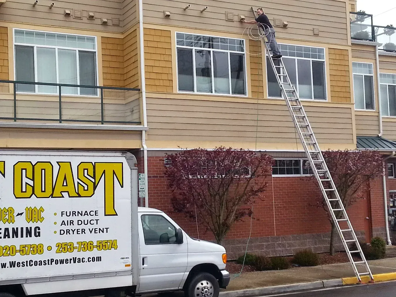 Man on ladder fixing building exterior, West Coast truck in front.