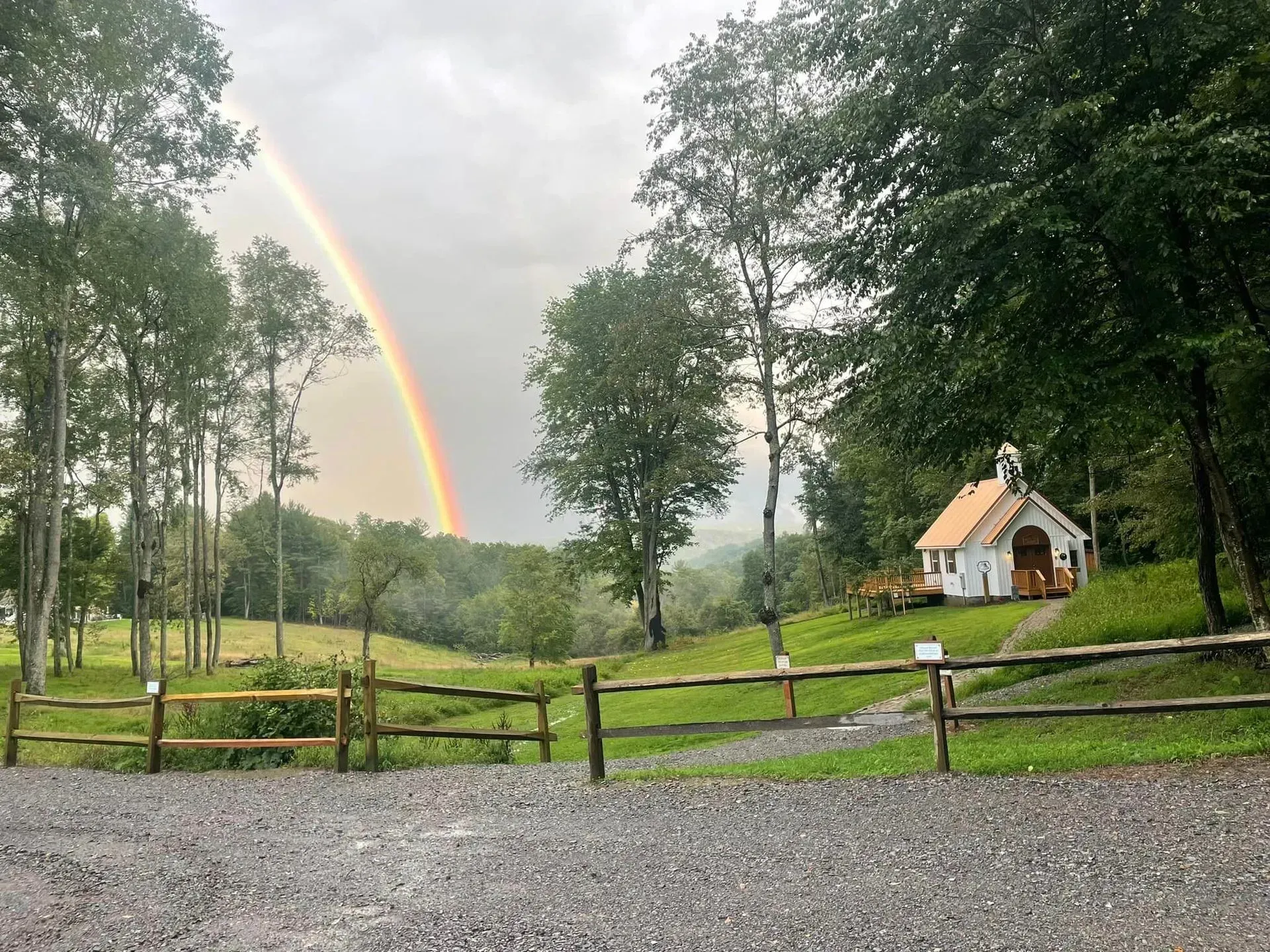 A white chapel with a small steeple sits on a grassy hill beside a split-rail fence under a bright rainbow.