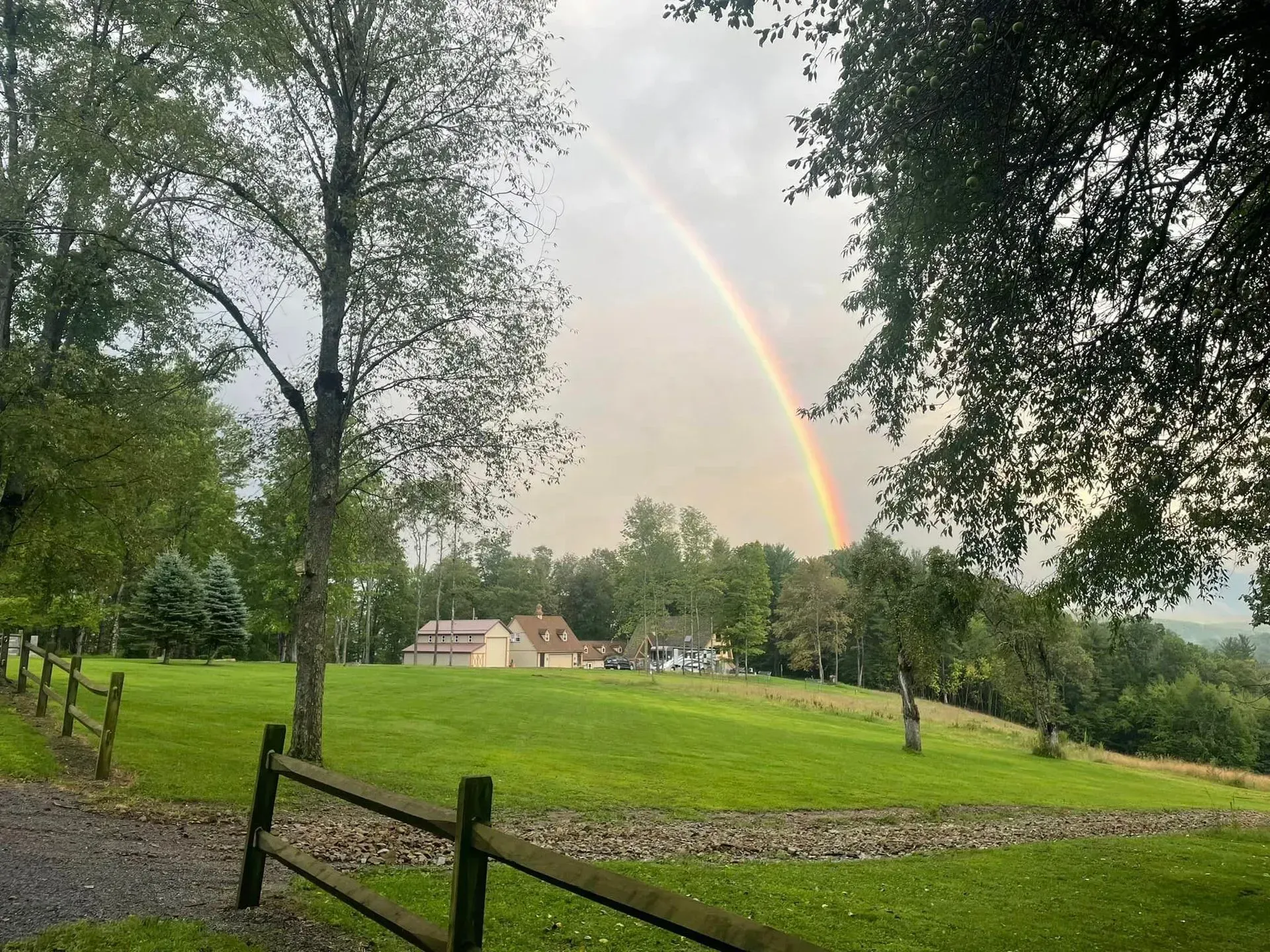 A bright rainbow arches over a green hillside with trees and houses in the distance under a cloudy sky.