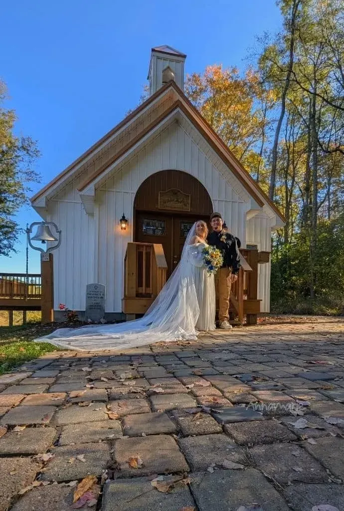A couple in wedding attire stands in front of a rustic white chapel on a sunny autumn day.