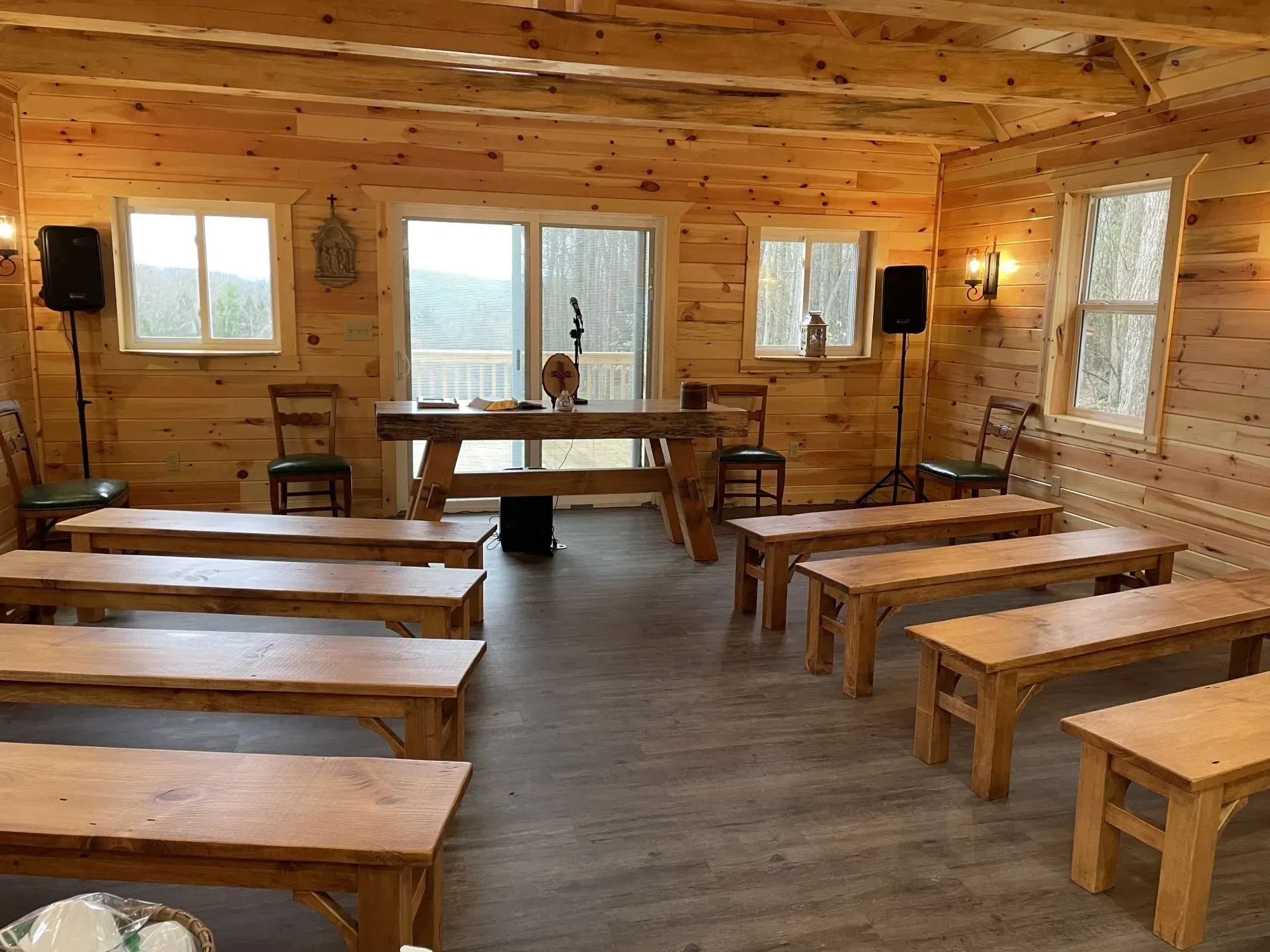 A rustic room with log walls, rows of wooden benches, a speaker podium, and windows overlooking a forest.