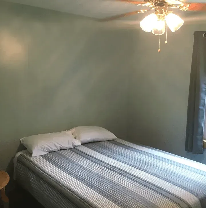 A view of a corner bedroom with light green walls, a bed with a grey and white striped quilt, and a lit ceiling fan.