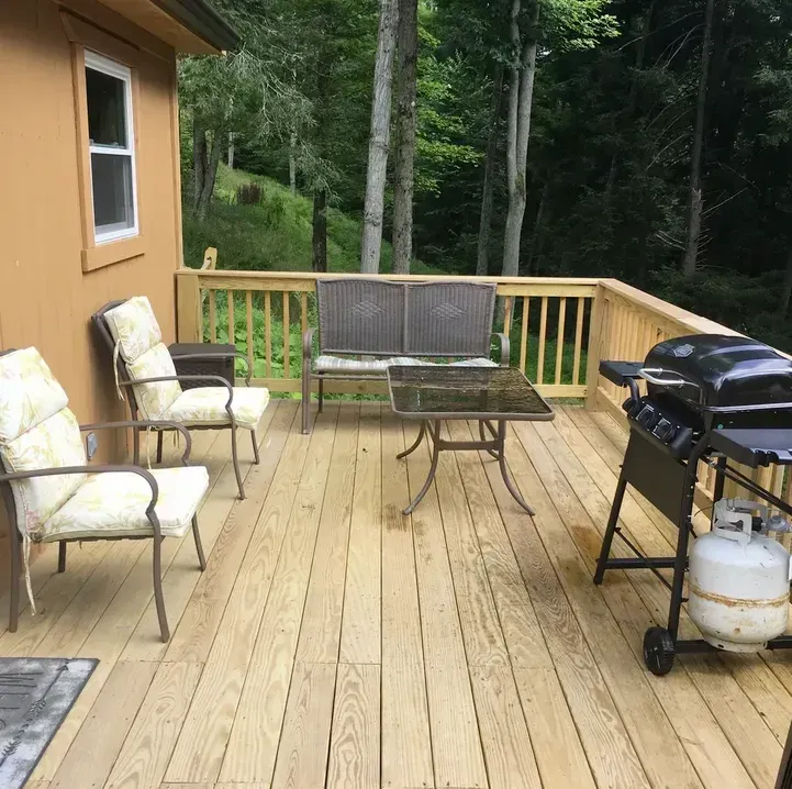 A wooden deck with two chairs, a bench, a small table, and a propane grill, overlooking trees.