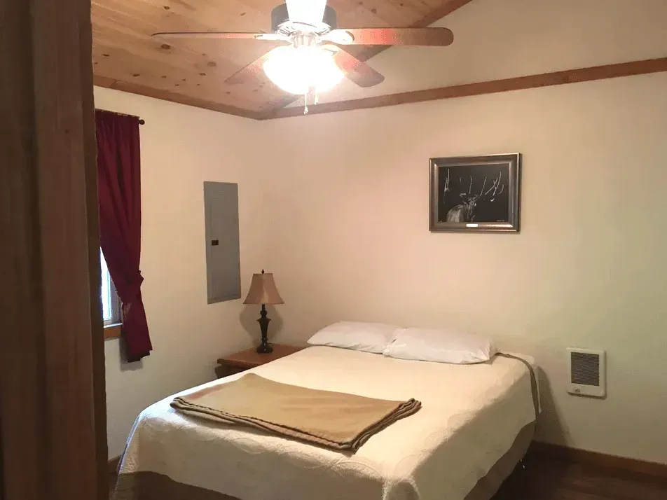 A bedroom with a queen bed, a lamp on a wooden nightstand, a framed picture on the wall, and a wood-paneled ceiling.