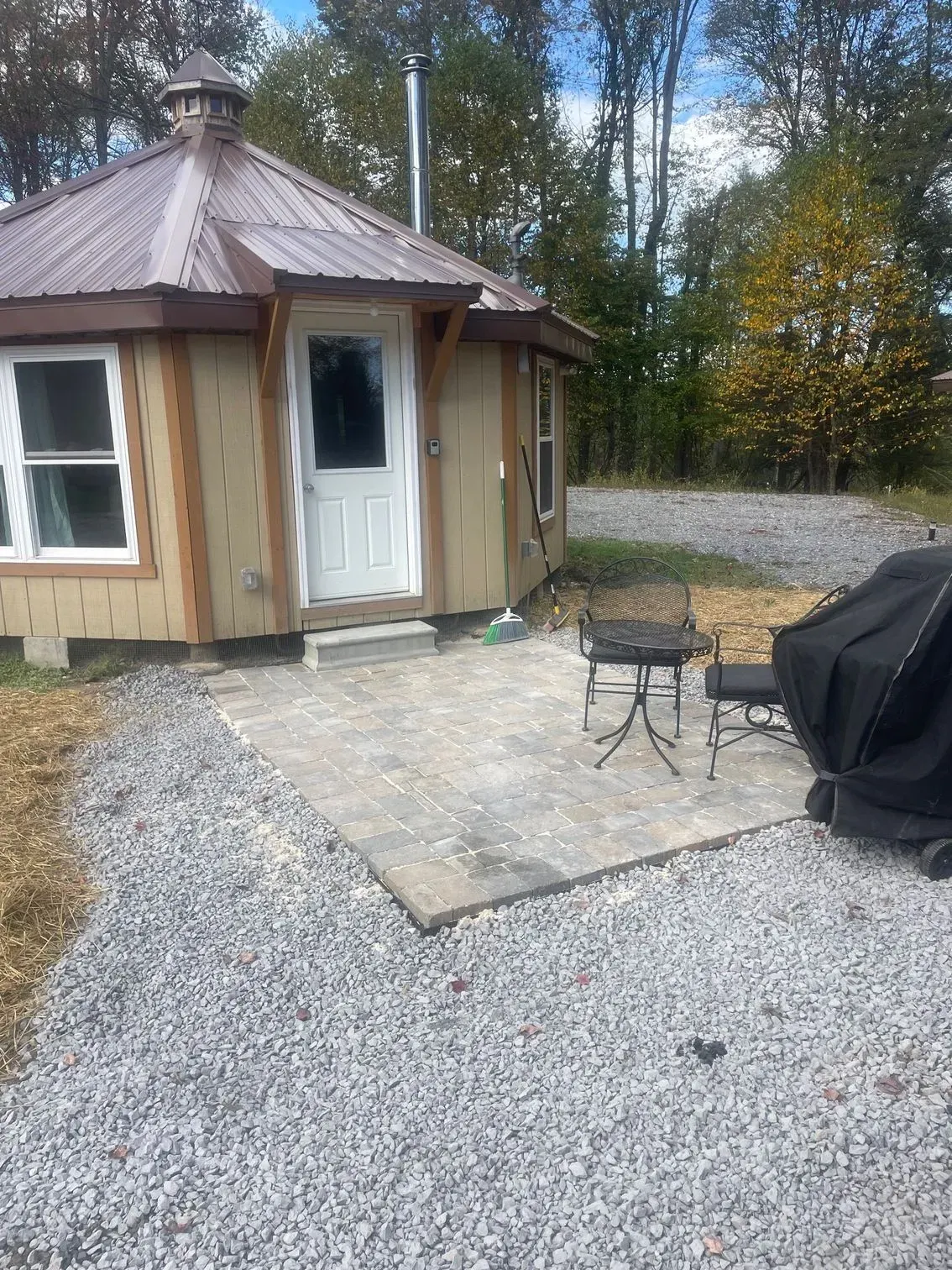 A yurt-style cabin with a brown metal roof, light wood siding, and a white door, set on a gravel lot with a paved patio.