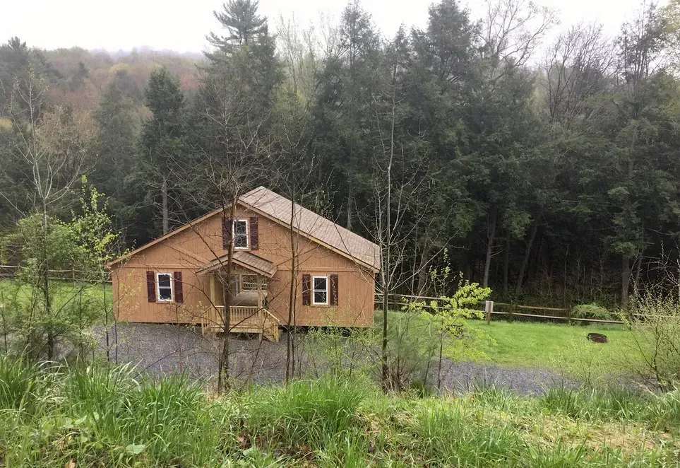A tan wooden house with dark red shutters, nestled in a lush, green woodland setting with a gravel driveway.