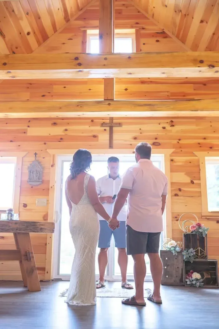 A couple holds hands before an officiant in a wooden chapel with a central cross and bright natural light.