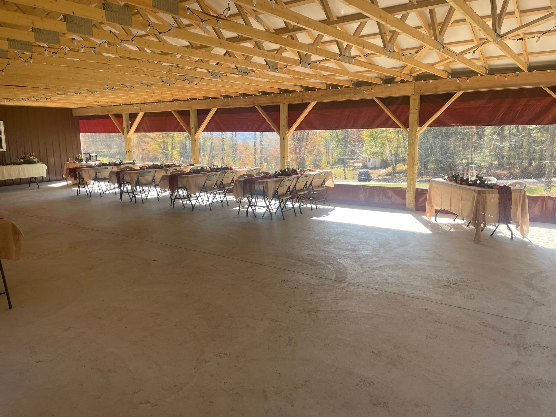 Large open pavilion with wooden beams, tables, and chairs on a concrete floor, lit by bright sunlight.