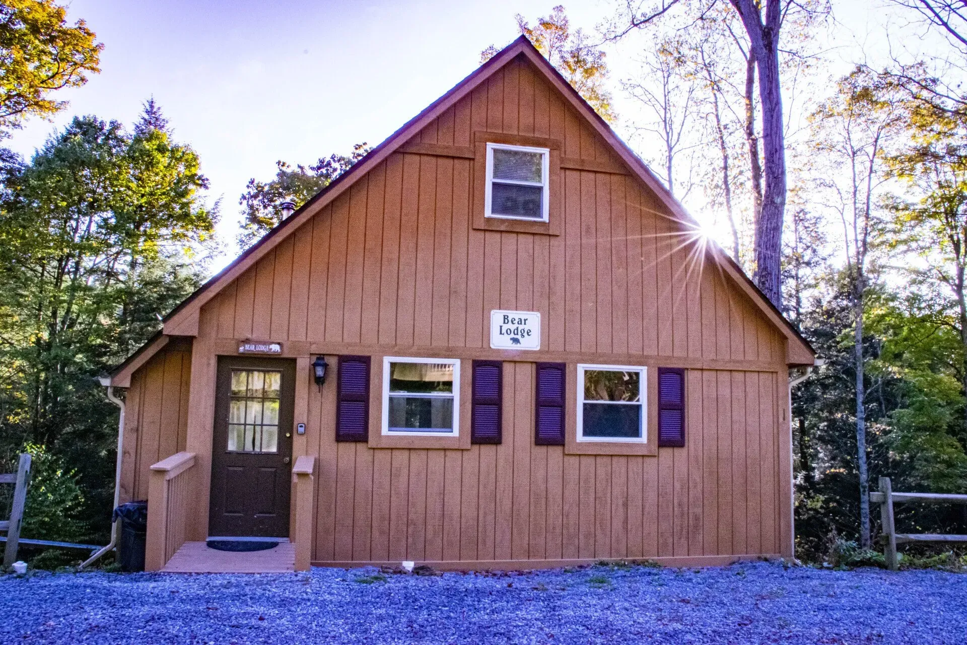 A brown wooden cottage with a gabled roof, two windows with shutters, and a front door, surrounded by trees.