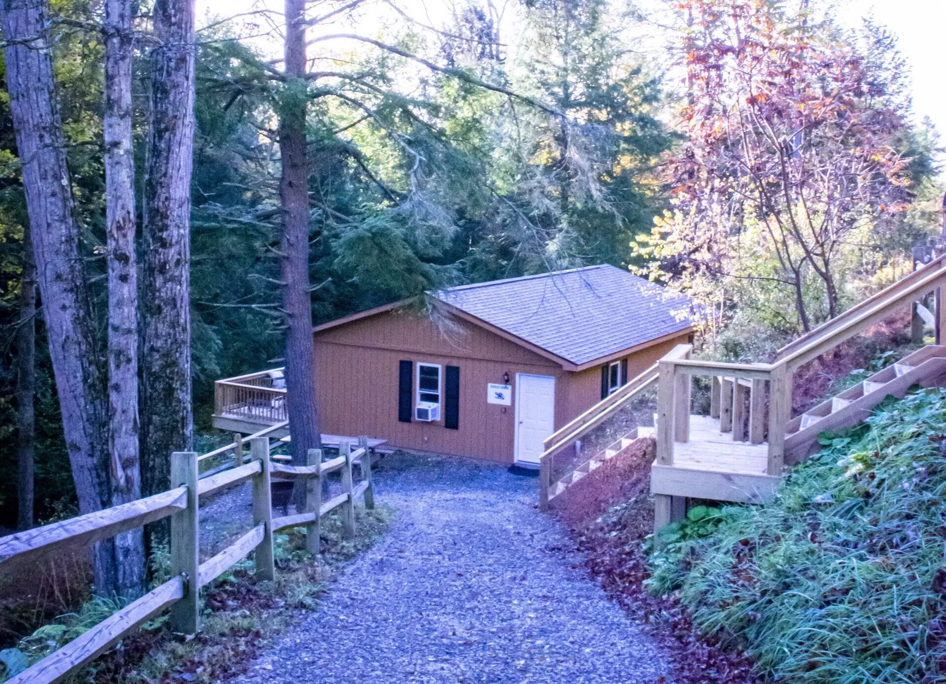 A gravel driveway leads to a brown cabin nestled in a forest, featuring a side deck and a wooden staircase.