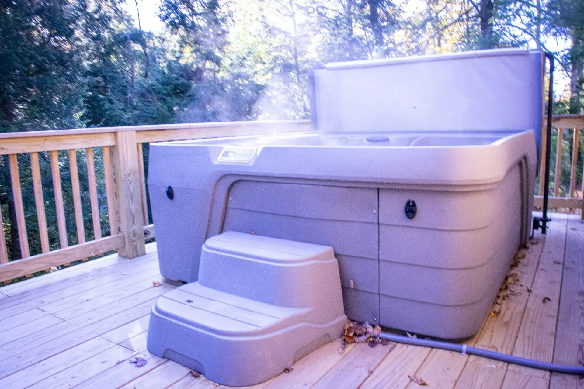 A grey, steaming hot tub with a matching step sits on a wooden deck surrounded by trees.