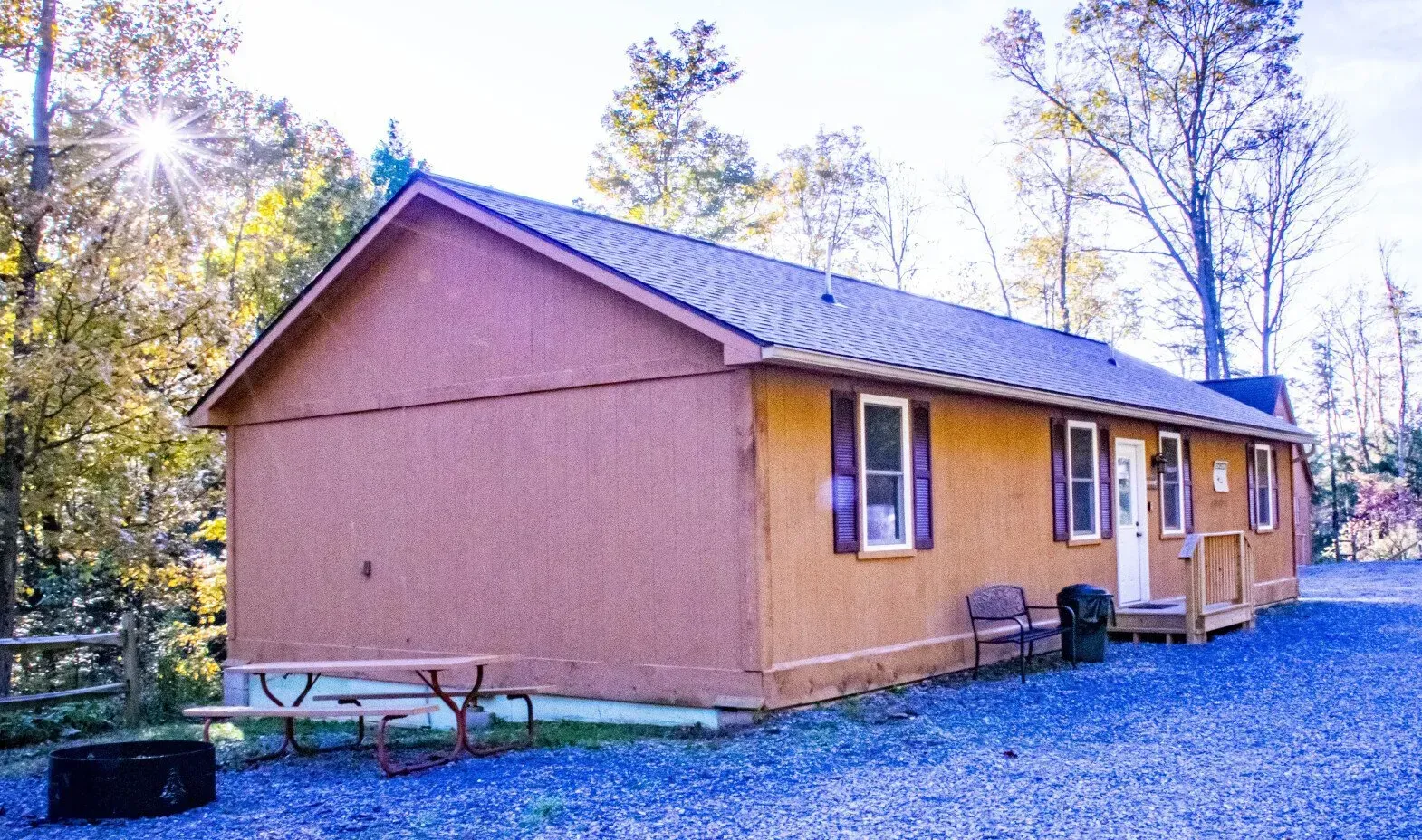 A tan cabin with a metal roof sits on a gravel lot next to a picnic table and a fire pit in a wooded setting.