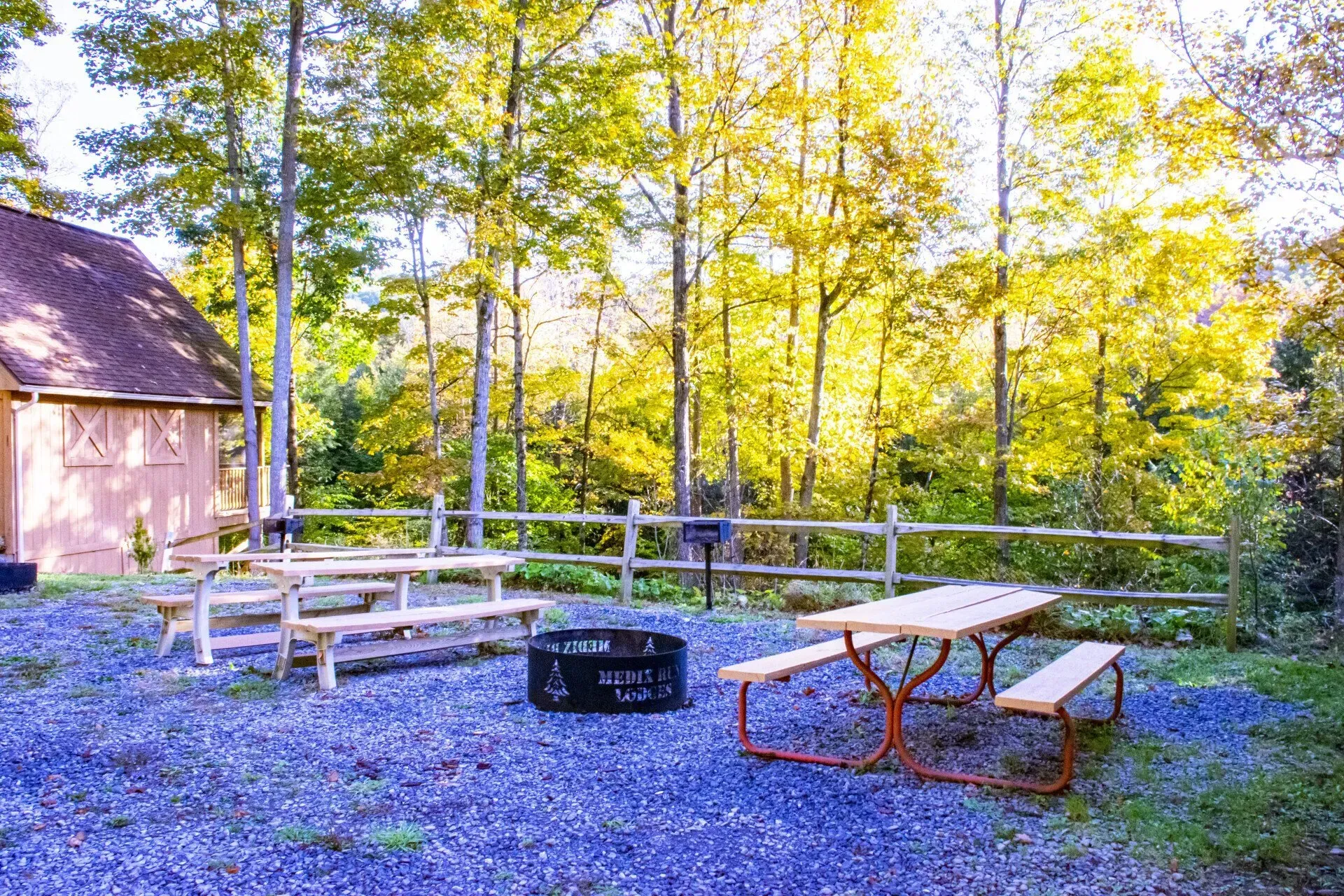 A gravel area with two wooden picnic tables and a fire pit, located in front of a building and autumn trees.
