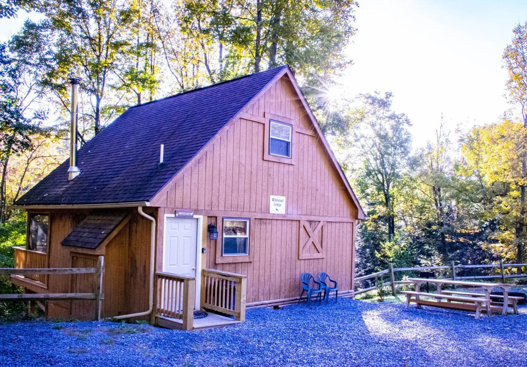 A tan, wooden A-frame cabin sits on a gravel lot near a picnic table with trees in the background under bright sunlight.