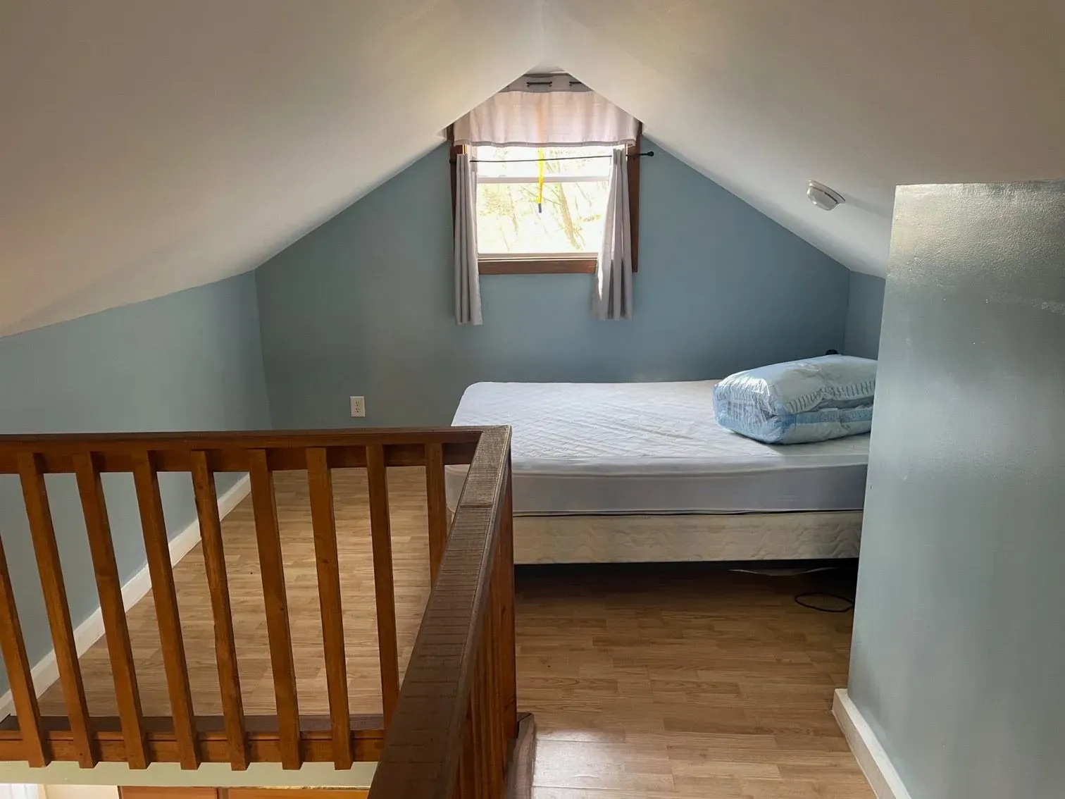 An attic room with blue walls, light-colored wood flooring, a bed, and a wooden railing overlooking a lower level.