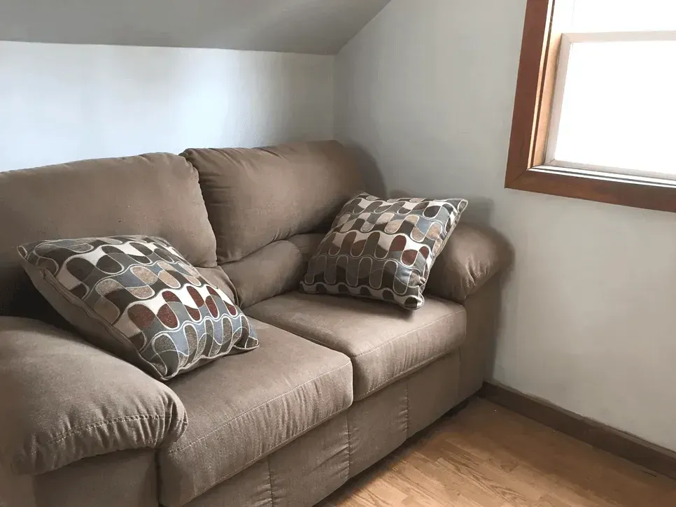 A brown loveseat with patterned accent pillows sits in a room with light walls and wood flooring near a window.