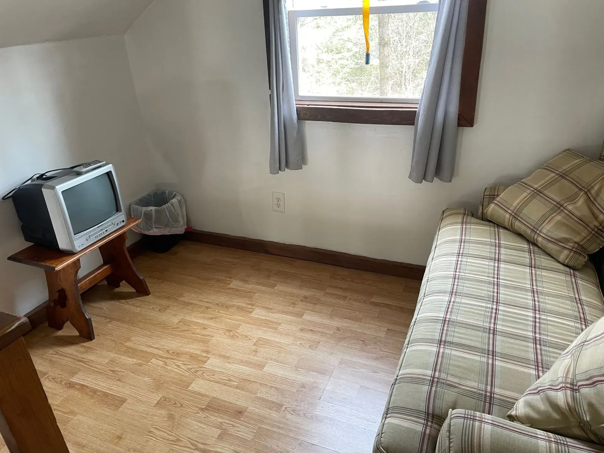 A small room with wood-patterned flooring, a plaid couch, a retro television on a wooden stand, and a window with curtains.