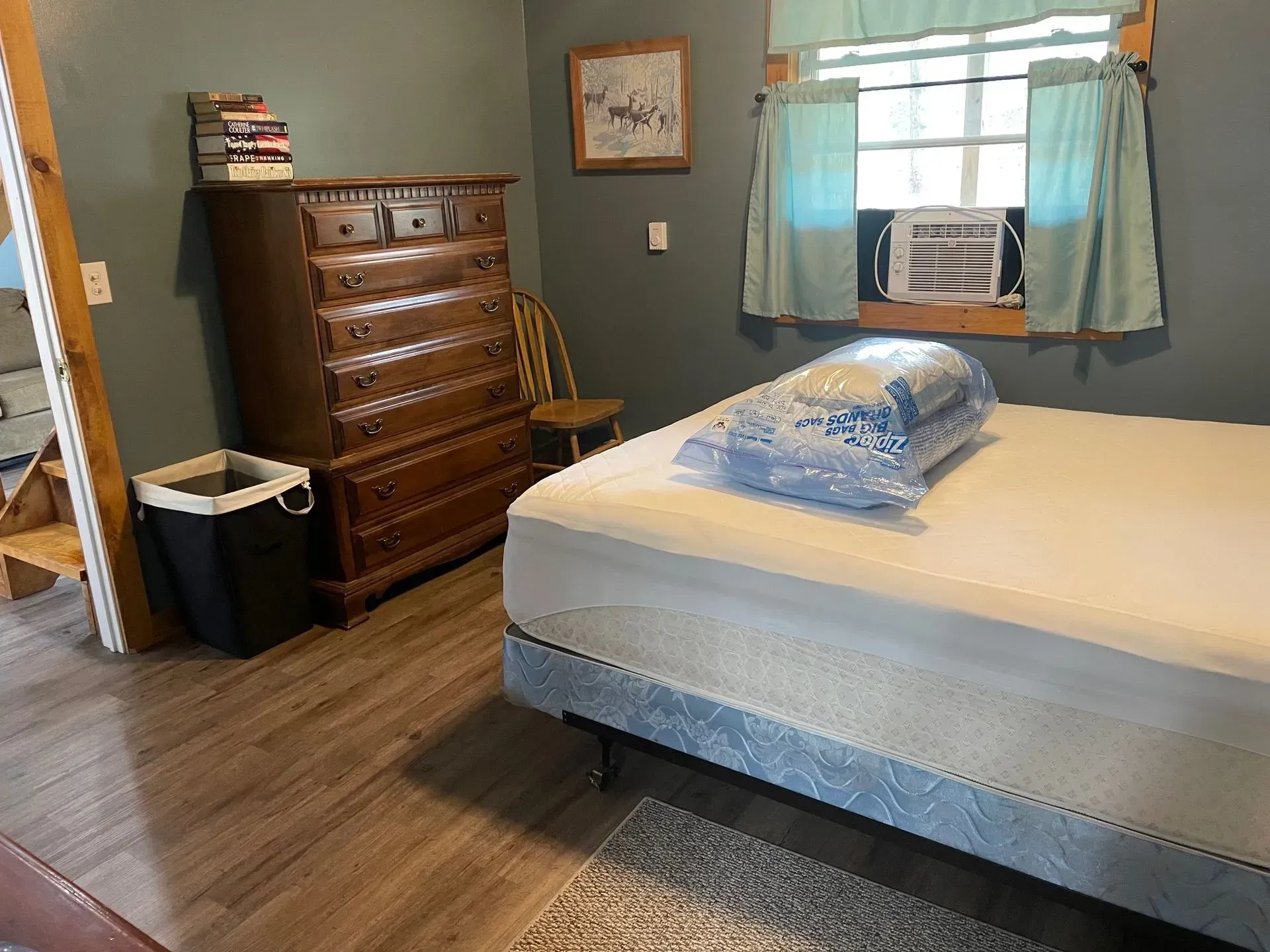 A bedroom featuring a bed with a bagged mattress topper, a wooden dresser, a chair, and a black laundry hamper on wood floors.