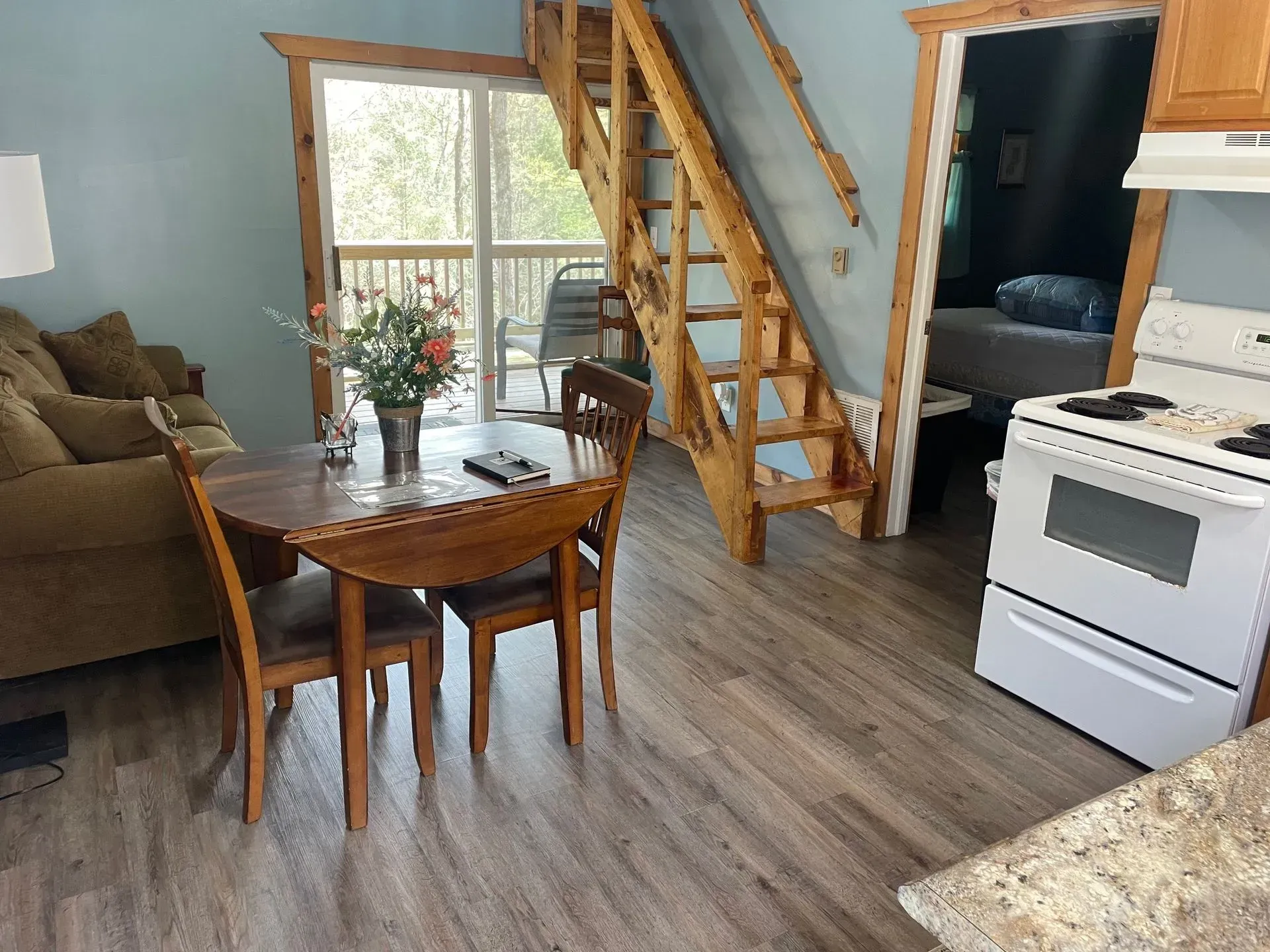 A small dining set sits in an open-concept room with a couch, a log staircase, and an adjacent kitchen.