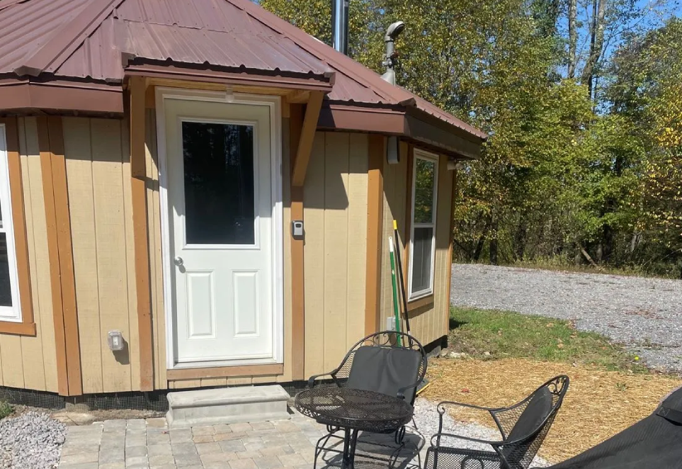 A tan yurt with a white door and metal roof, featuring a small stone patio with two black metal chairs and a table.