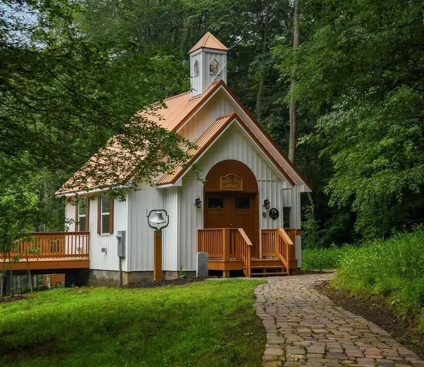 A small, white wooden chapel with a copper roof and steeple sits in a lush, green forest at the end of a stone path.