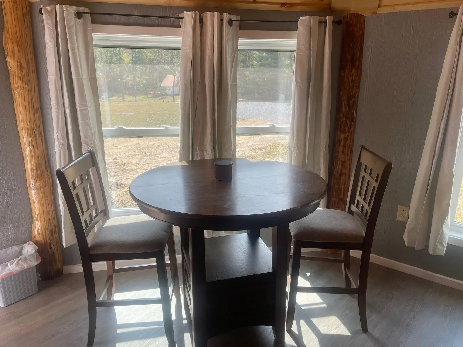 A round dark wood pub-style table with two matching chairs placed before a large window with white curtains.