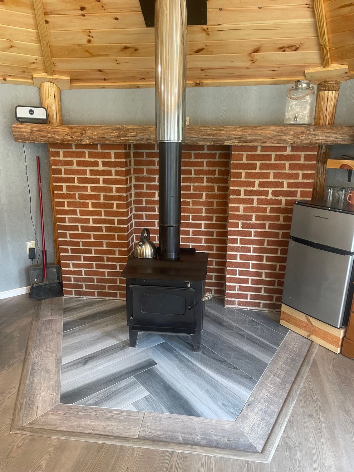 Wood stove with silver chimney in a small brick-lined room with a chevron wood floor