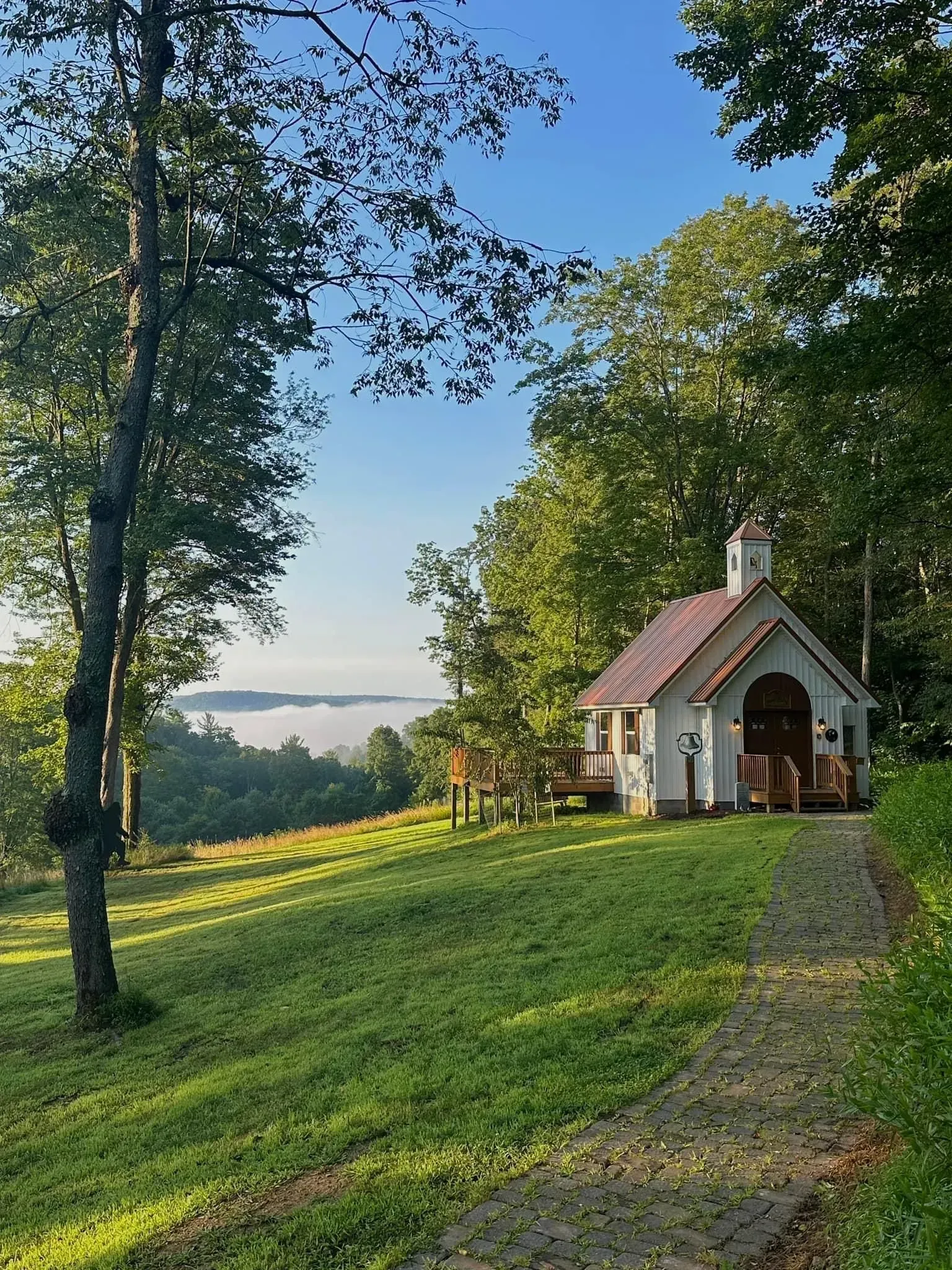A small white chapel with a brown roof sits on a green hillside overlooking a misty valley under a clear blue sky.