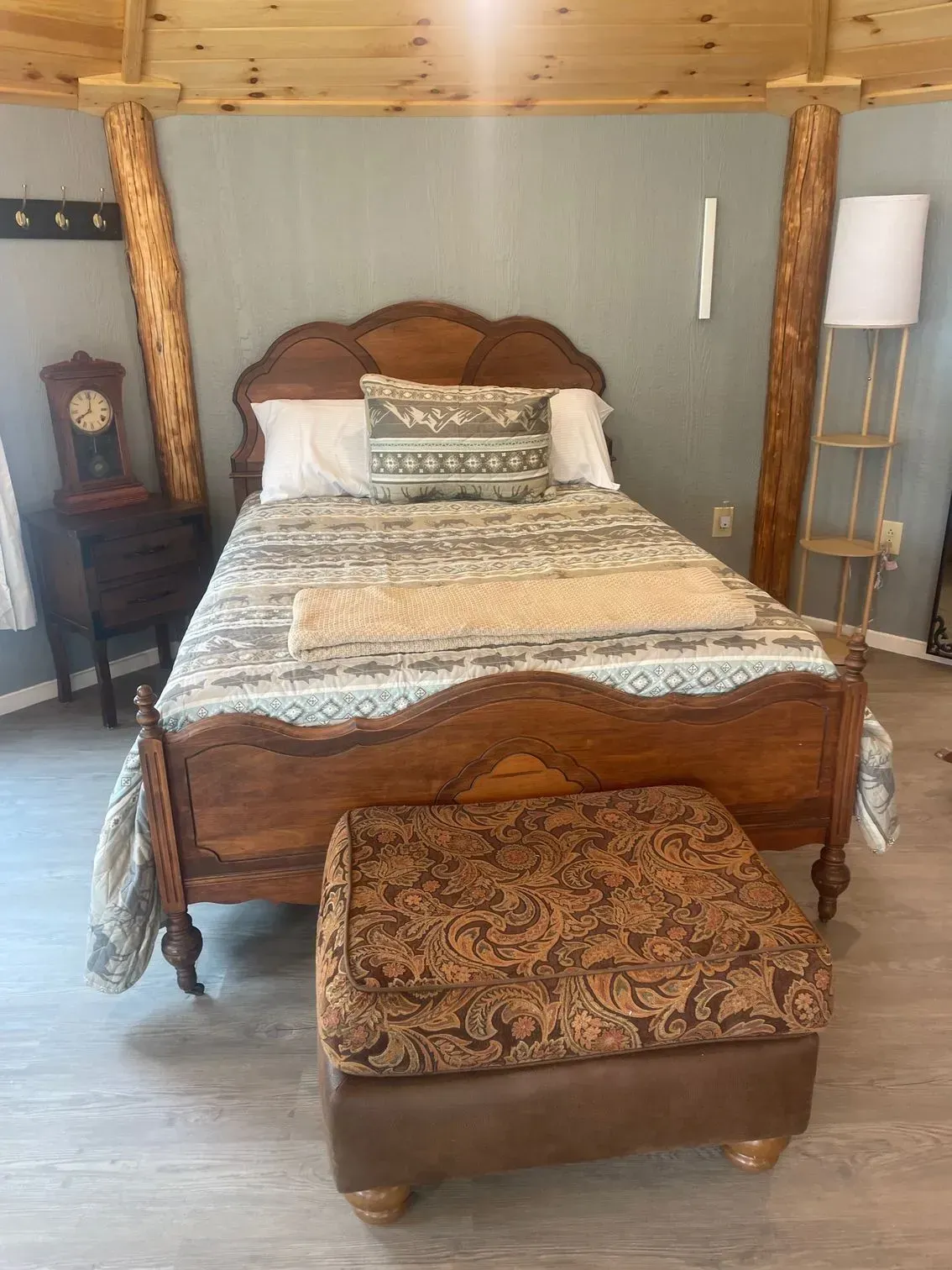 A rustic bedroom with a wooden bed frame, patterned bedding, a dark wooden nightstand with a clock, and a storage ottoman.