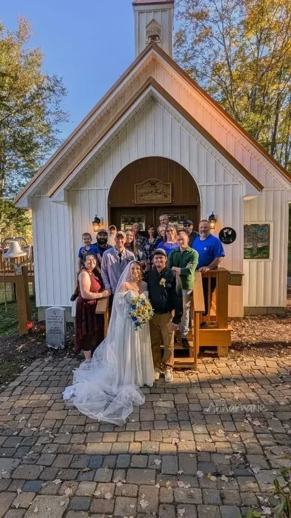 A bride and groom pose with their wedding guests in front of a white wooden chapel on a sunny day.