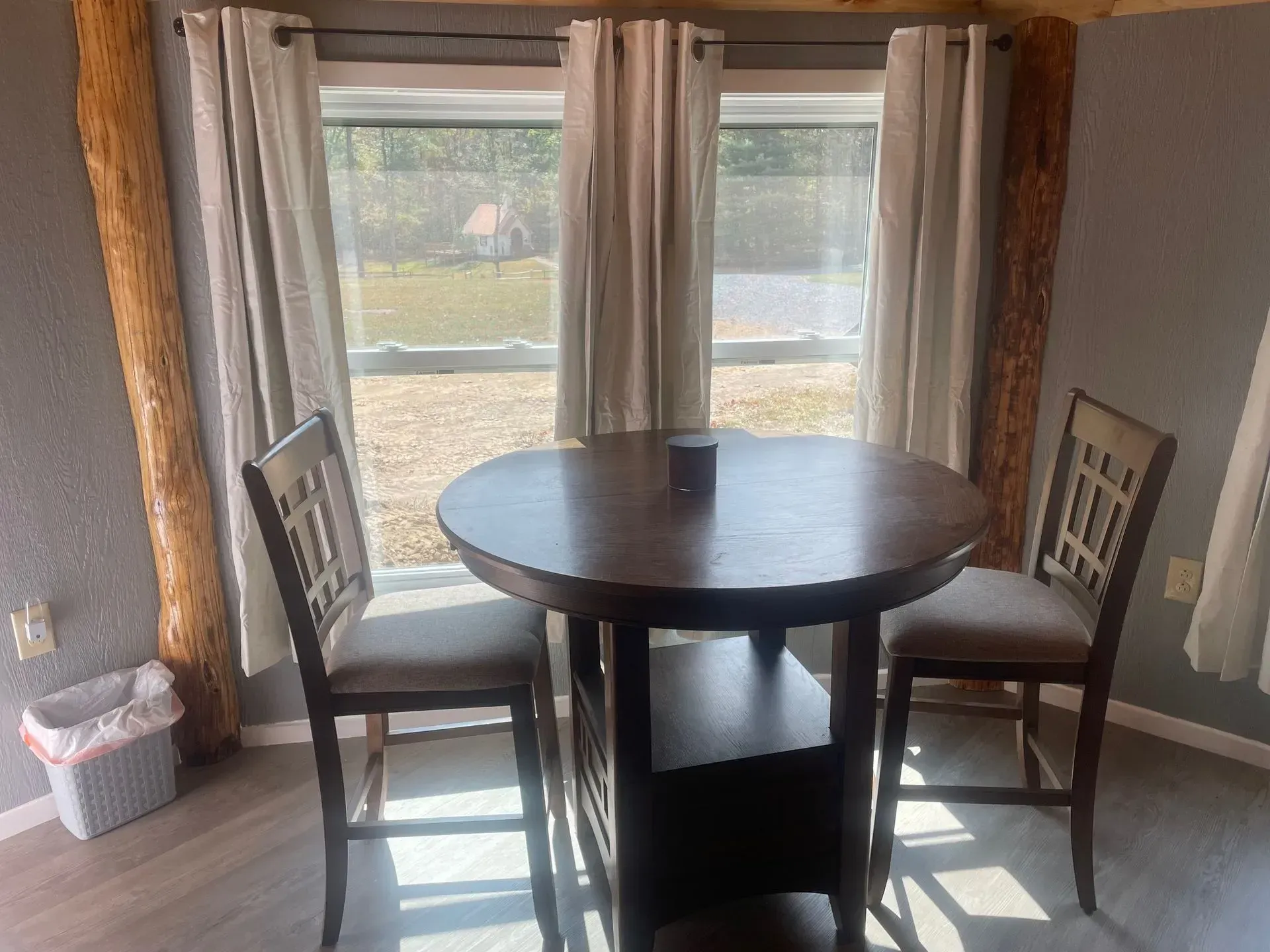 A round, dark wood table with a storage base and two high-backed chairs positioned in front of a window with curtains.